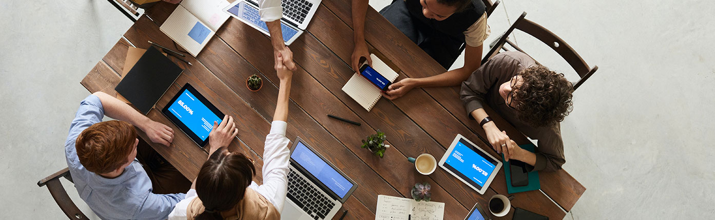 Two people shake hands across a wooden table while three others sit nearby using mobile devices.