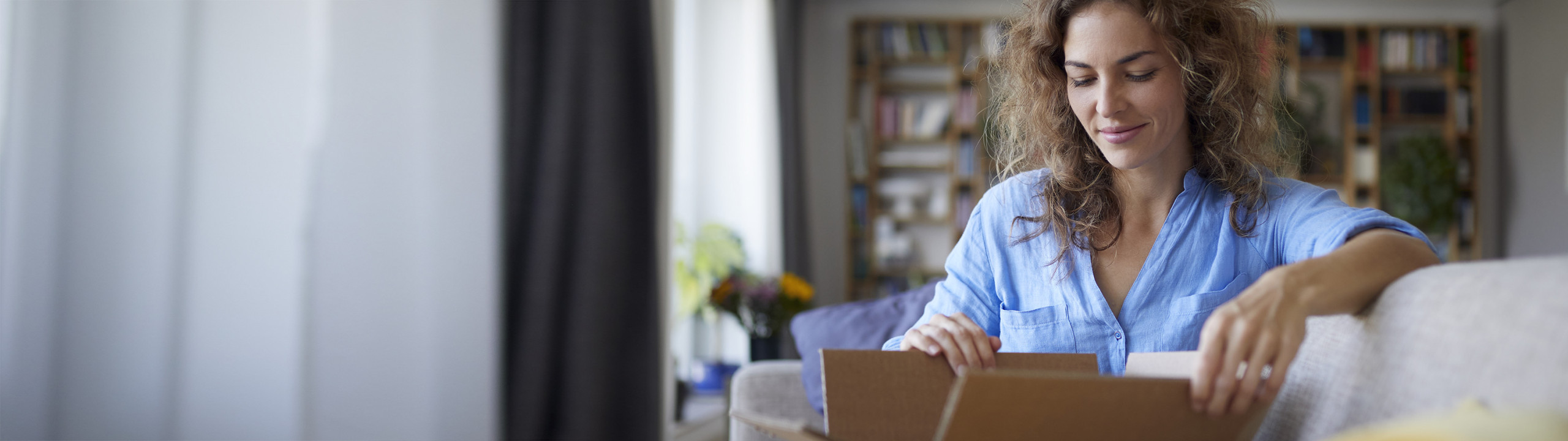 A brunette woman sitting on a couch in a warm living room, opening a box with anticipation.