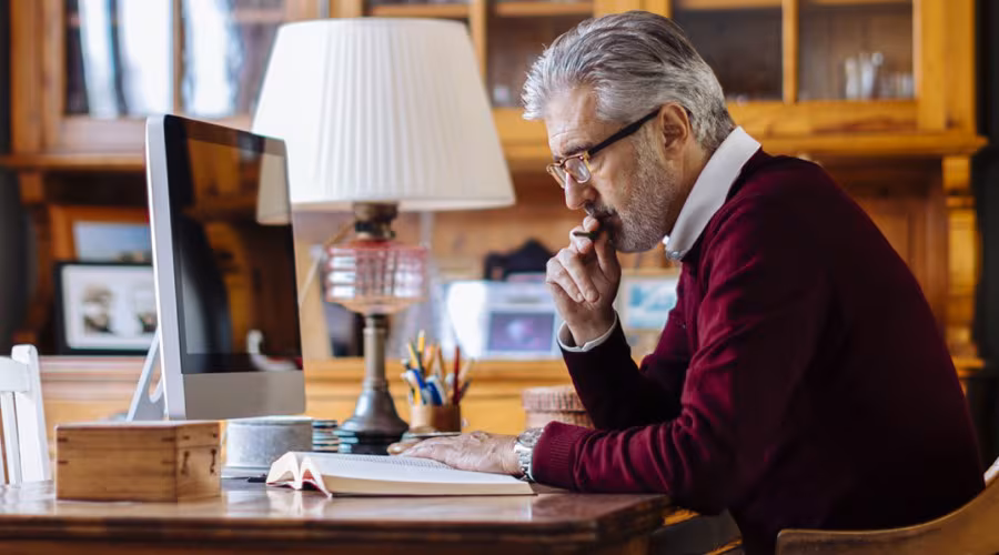 man-home-computer An older gentleman in a burgundy sweater sits at a wooden desk, intently reading a book in a warmly furnished office.